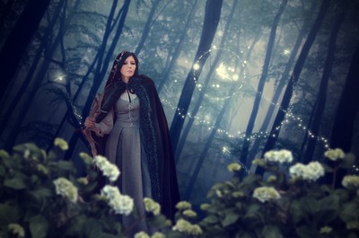 The image shows an individual with an obscured face standing in a misty forest, holding a bow. They are wearing a long cloak and a dress, surrounded by white flowers in the foreground. Sunlight filters through the dense trees in the background, creating an ethereal atmosphere. It suggests themes of fantasy and adventure.