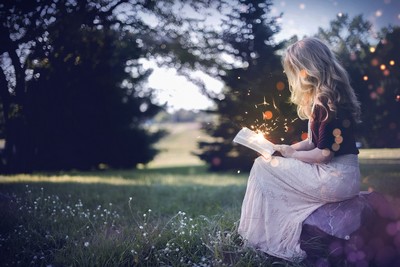 A person sitting in a grassy field during twilight, reading a glowing book with small light particles floating around, creating a magical atmosphere. It seems to evoke a sense of wonder and the magical world of fairy tales or fantasy stories.