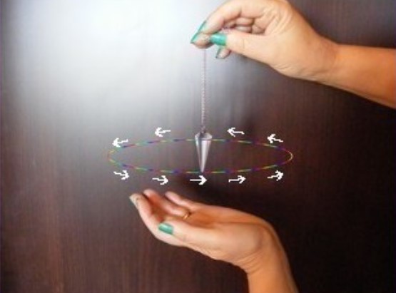 A pair of hands holding a pendulum in motion. The top hand holds the chain, while the bottom hand is open beneath it. Arrows indicate the circular path of the pendulum's swing, illustrating the direction of its movement. The background is a dark, smooth surface.